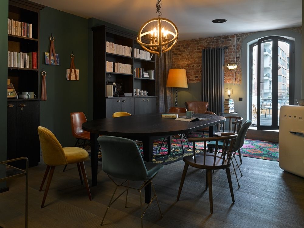 A meeting room featuring a round table, colorful chairs, bookshelves, and large windows overlooking an outdoor space.