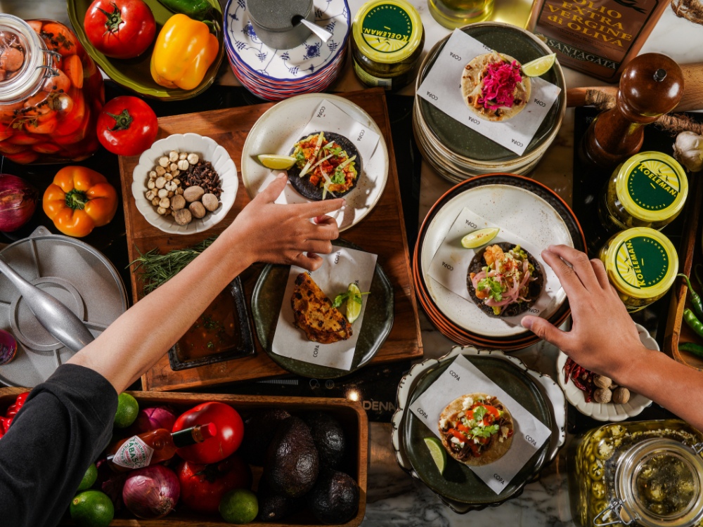 A vibrant spread of colorful tacos and fresh ingredients, with hands reaching for delicious dishes amidst a lively kitchen scene.